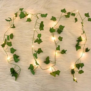 May include: Artificial ivy garland with small, warm white LED lights. The green leaves are arranged along a thin, flexible wire, creating a decorative vine. The lights are evenly spaced, providing a soft glow. Ideal for home decor and event decorations.