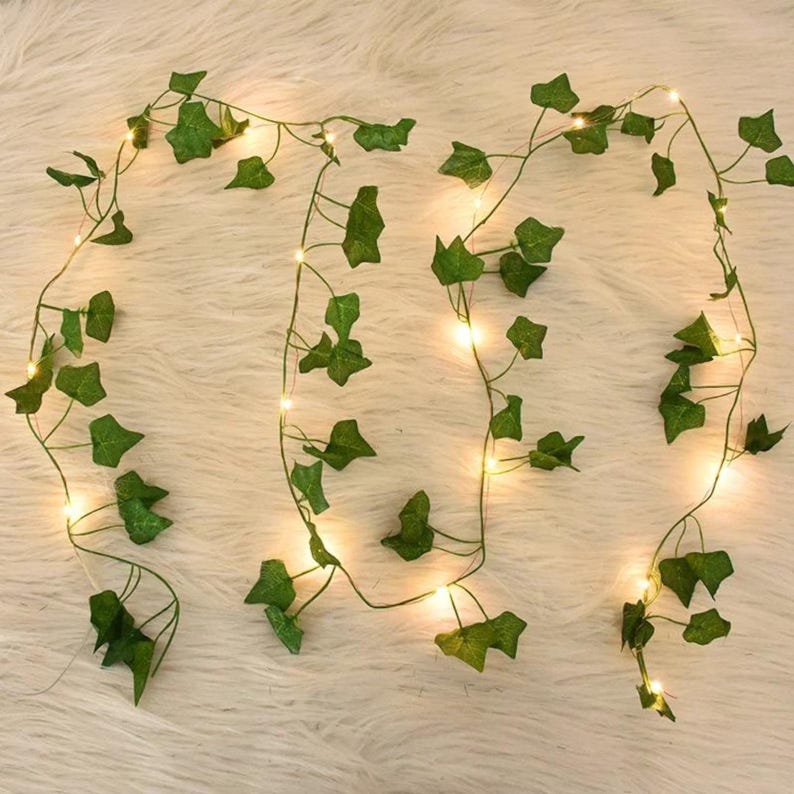 May include: Artificial ivy garland with small, warm white LED lights. The green leaves are arranged along a thin, flexible wire, creating a decorative vine. The lights are evenly spaced, providing a soft glow. Ideal for home decor and event decorations.
