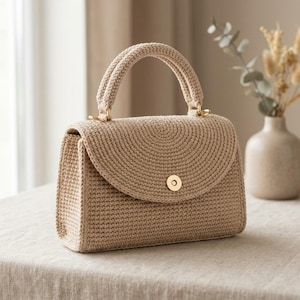 May include: A beige crochet handbag featuring a structured rectangular body, a semi-circular flap, and a single top handle. The bag is secured with a gold-toned circular magnetic clasp and is crafted from a dense, uniform yarn stitch pattern.