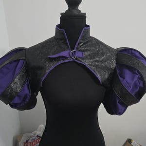 Könnte beinhalten: Schwarze und lila Bolerojacke mit hohem Kragen und Puffärmeln. Die Jacke besteht aus strukturiertem schwarzem Material mit lila Akzenten und einem dekorativen Verschluss. Das Kleidungsstück wird auf einer schwarzen Schaufensterpuppe präsentiert.