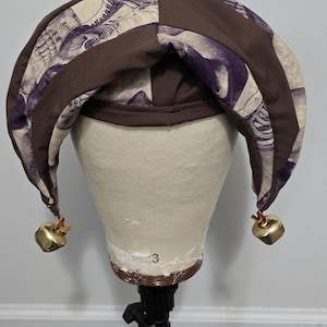 Peut inclure: Un chapeau de bouffon marron et violet avec un motif et des cloches dorées. Le chapeau est présenté sur une tête de mannequin. Le tissu du chapeau a un fond crème avec des illustrations violettes.