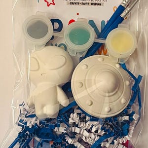 May include: A clear plastic bag containing a Kulay at Guhit plaster figurine art kit. The kit includes two white plaster figures, one shaped like an alien and one like a flying saucer, three small paint pots, and two paintbrushes on blue shredded paper.