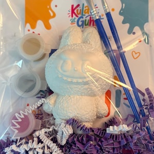 May include: A craft kit in a clear plastic bag containing a white textured rabbit figurine, two blue-handled paintbrushes, three small paint pots, and purple paper filler. A card with the text Kula at Guhit is visible in the background.