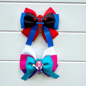 May include: Two layered hair bows made of grosgrain ribbon. The top bow features red, blue, and black ribbons with a Spider-Man character charm. The bottom bow features white, pink, purple, and teal ribbons with a Spider-Gwen character charm.