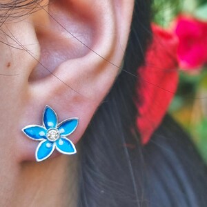 Pode incluir: Um brinco em forma de flor de cinco pétalas com esmalte azul brilhante preenchendo cada pétala e uma única pedra preciosa redonda e transparente cravada no centro. O brinco tem uma borda metálica prateada que contorna as pétalas e o centro.