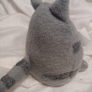 May include: Gray plush cat toy with a striped tail. The cat is sitting on a white surface.