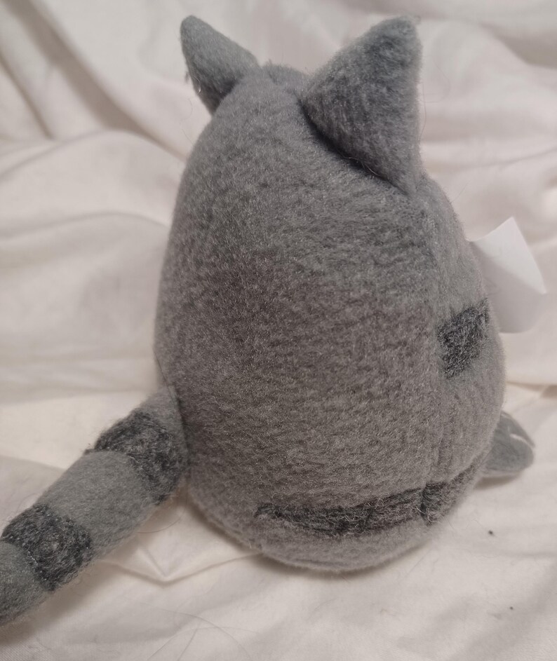 May include: Gray plush cat toy with a striped tail. The cat is sitting on a white surface.