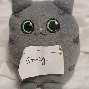 May include: Gray plush cat toy with large green eyes and a pink nose. The toy is sitting on a white surface with a note that says "Stacey".