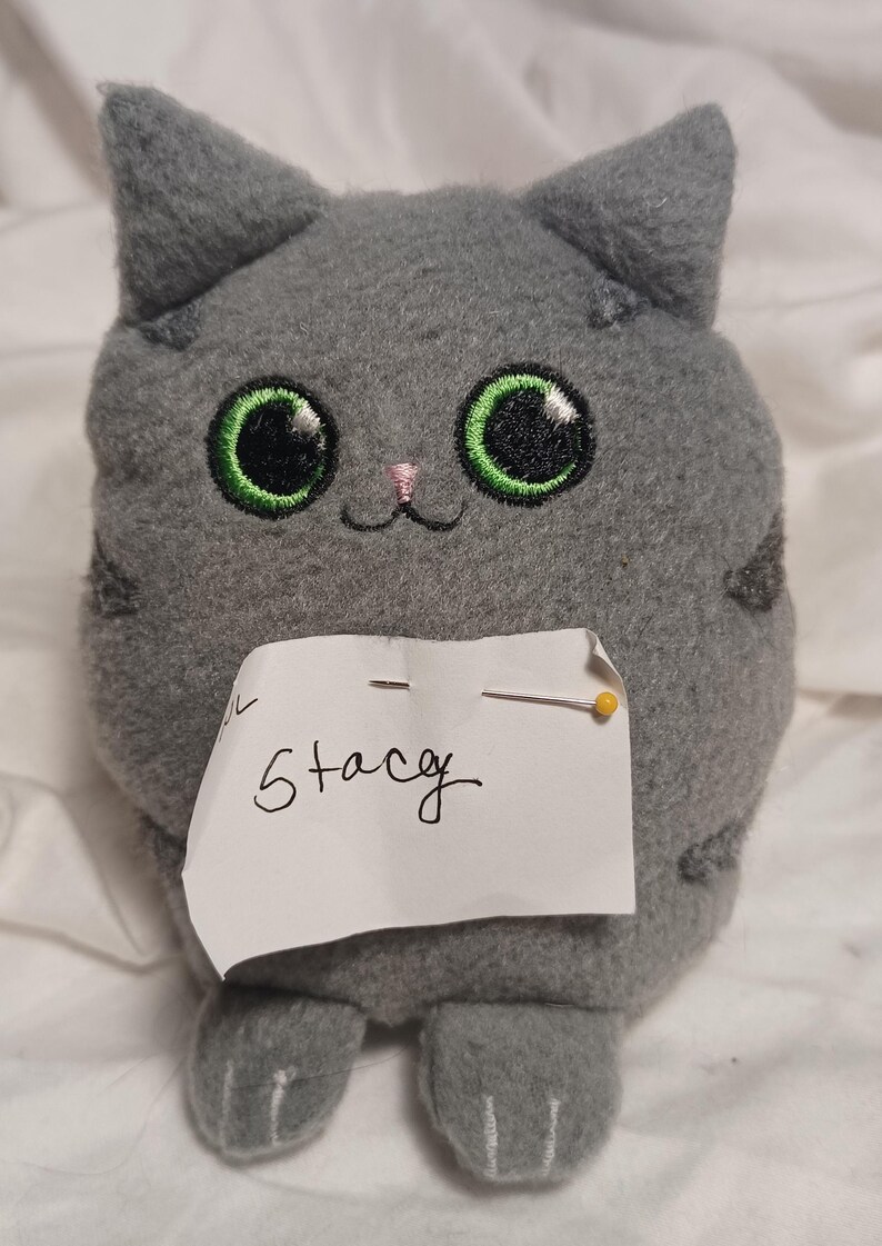 May include: Gray plush cat toy with large green eyes and a pink nose. The toy is sitting on a white surface with a note that says "Stacey".
