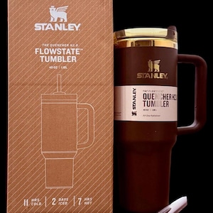 Puede incluir: Un vaso Stanley Flowstate marr&oacute;n con asa y borde dorado, junto a su caja de cart&oacute;n. La caja presenta el logotipo de Stanley y detalles del producto, incluyendo "40 OZ | 1.18L". El vaso est&aacute; dise&ntilde;ado para la hidrataci&oacute;n durante todo el d&iacute;a.