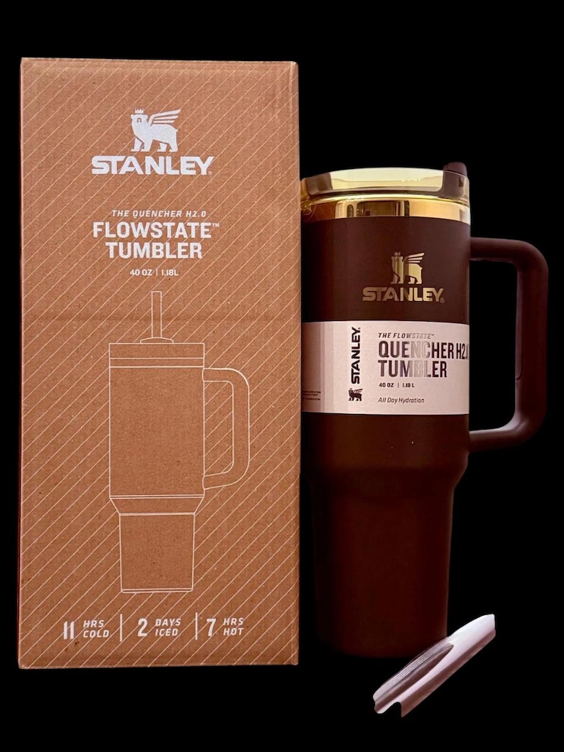 Puede incluir: Un vaso Stanley Flowstate marr&oacute;n con asa y borde dorado, junto a su caja de cart&oacute;n. La caja presenta el logotipo de Stanley y detalles del producto, incluyendo "40 OZ | 1.18L". El vaso est&aacute; dise&ntilde;ado para la hidrataci&oacute;n durante todo el d&iacute;a.