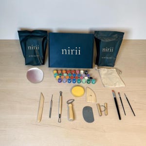 May include: A complete air dry clay kit with a dark blue box and bags labeled "nirii". The kit includes clay, paints, tools, and brushes. The paints are in small, colorful containers. A beige drawstring bag is also included.
