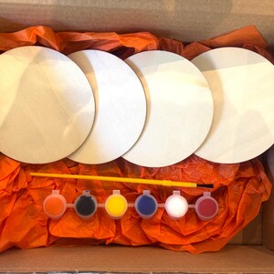 Pode incluir: Um kit de artesanato numa caixa de cartão, com quatro círculos de madeira inacabados, potes de tinta de várias cores e um pincel amarelo. O kit está aninhado em papel de seda laranja.