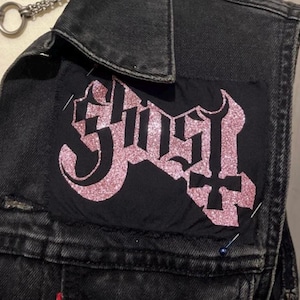 Op de afbeelding: Een zwarte spijkerweste met een roze glitter patch met de tekst "Ghost" in een gotisch lettertype. De patch is met spelden op de vest genaaid. Een zilveren ketting is zichtbaar aan de bovenkant.