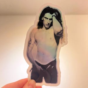 May include: Holographic sticker showing a shirtless person with long, dark hair, and black trousers. The person is touching their hair with one hand. The sticker has a rainbow sheen and is held by a hand.