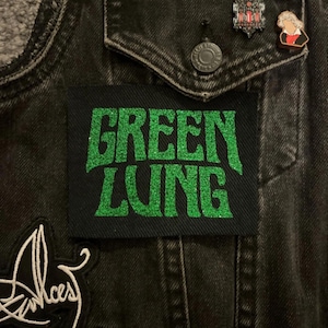 Op de afbeelding: Zwarte denim jas met een "GREEN LUNG" patch in groene glitterletters. De jas heeft zilveren studs op de schouders en twee spelden op de zak. Een wit en zwarte patch is zichtbaar links onder.