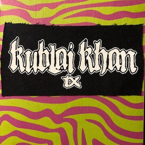 Puede incluir: Un parche negro con el texto blanco "Kublai Khan" en una fuente estilizada. Debajo del texto hay un pequeño emblema blanco. El fondo presenta un patrón de rayas de cebra rosas y amarillo-verdes.