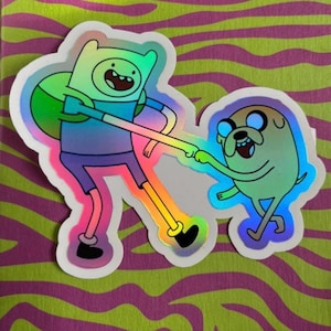 May include: Holographic sticker of Finn and Jake from Adventure Time. Finn wears a blue shirt and purple shorts, Jake is a yellow dog. The sticker has a white border and is on a green and purple striped background.