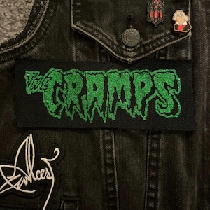 May include: Black denim jacket with a rectangular patch featuring the band name "The Cramps" in green glitter lettering. The jacket also has several pins and patches.