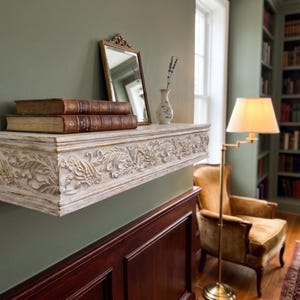 May include: A white and gold carved wooden shelf with two leather-bound books, a small mirror, and a vase with dried flowers. The shelf is mounted on a green wall, creating a classic, elegant display.
