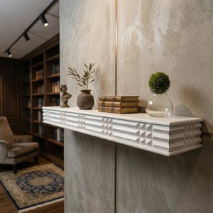 May include: A white, geometric-patterned shelf is mounted on a textured wall, displaying decorative items. These include a small bust, a vase with olive branches, stacked books, and a glass vase with a green plant. The shelf is a focal point.