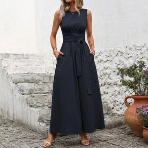 May include: A sleeveless, navy blue dress with a wrap-around waist and a matching belt. The dress has a full skirt with pockets and is worn with sandals. The background features a white wall and stone steps.