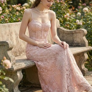 May include: A light pink lace dress with spaghetti straps. The dress features a fitted bodice and a flowing skirt, with floral lace detailing throughout. The dress is worn outdoors, with a garden in the background.