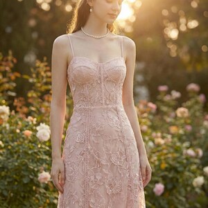 May include: A light pink, floor-length dress with a floral lace overlay. The dress features a fitted bodice with spaghetti straps and a sweetheart neckline. The dress is worn outdoors in a garden setting.