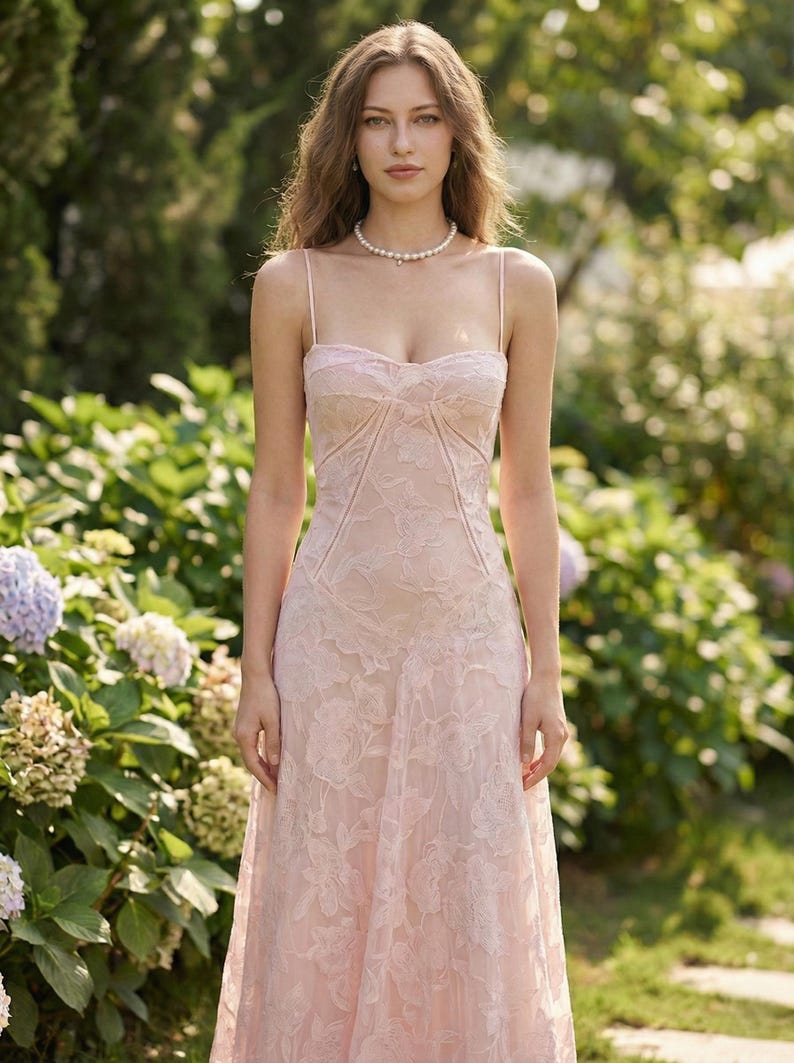 May include: A light pink, floor-length dress with a floral lace overlay. The dress features spaghetti straps and a fitted bodice with a sweetheart neckline. A pearl necklace is worn. The background includes green foliage and flowers.