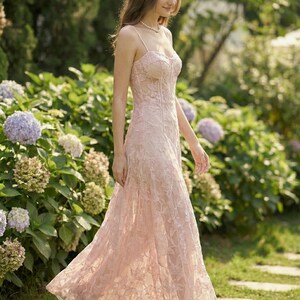 May include: A light pink, floor-length formal dress with a floral lace overlay. The dress features a fitted bodice with spaghetti straps and a flowing skirt. The image is set outdoors with a garden background.