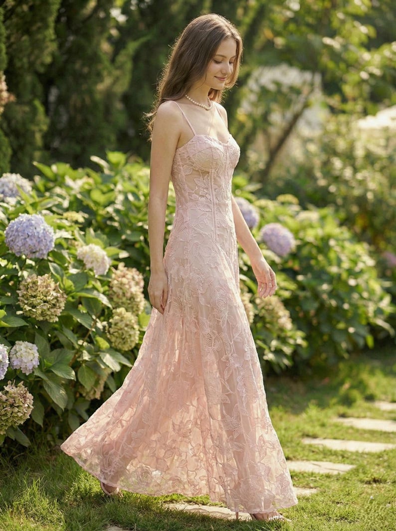 May include: A light pink, floor-length formal dress with a floral lace overlay. The dress features a fitted bodice with spaghetti straps and a flowing skirt. The image is set outdoors with a garden background.