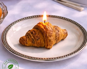 May include: A croissant-shaped candle with a lit wick sits on a white plate with a silver beaded rim. The candle is a golden brown color, and the flame is bright yellow. The plate is on a white tablecloth. An "Eco Product" stamp is visible.