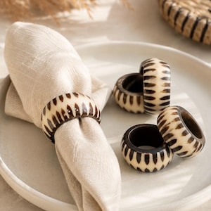 May include: A beige linen napkin is held by a decorative napkin ring. The ring is cream-colored with a dark brown pattern. Several other rings are on a white plate, alongside a woven basket.