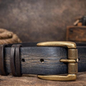 Può includere: Primo piano di una cintura in pelle nera con fibbia in ottone. La cintura ha un aspetto invecchiato ed è fissata con due passanti in pelle. La fibbia è un design classico con una finitura strutturata. La cintura è su una superficie di legno.