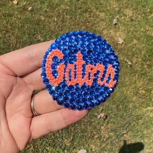 May include: A circular emblem covered in dark blue rhinestones featuring the word Gators spelled out in orange rhinestones across the center. The surface is densely packed with reflective, faceted stones that create a textured, sparkling appearance.