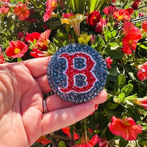 May include: A round, bedazzled emblem featuring the letter "B" in red, surrounded by a blue and white border. The emblem is covered in small, sparkling crystals. The background includes vibrant red and orange flowers.