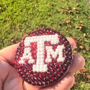May include: A circular phone grip featuring the Texas A&M University ATM logo in white, surrounded by a dense arrangement of small, reflective dark red rhinestones that cover the entire surface of the round accessory.