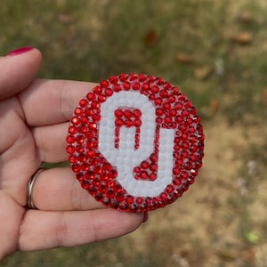 May include: A circular emblem featuring the University of Oklahoma logo, crafted with red and white rhinestones. The design displays a white script OU monogram set against a background of red crystals, creating a textured, sparkling appearance.