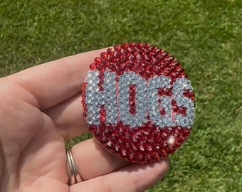 May include: A circular phone grip accessory featuring a red background made of small, reflective rhinestones. The word HOGS is spelled out in the center using clear, sparkling rhinestones that contrast against the red circular base.