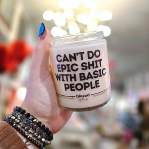 May include: A clear glass jar candle with a white wax interior. The candle has black text that reads "CAN'T DO EPIC SHIT WITH BASIC PEOPLE." The brand name "blazed" is printed below the text. The candle is held in a hand.