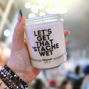 May include: A white soy candle in a clear glass jar with the text "LET'S GET THAT 'STACHE WET" in black. The candle is held in a hand with a bracelet. The candle is from "blazed" and says "lighten up".
