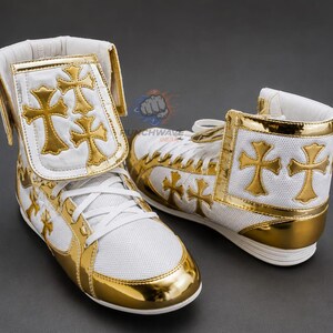 Puede incluir: Un par de botas de boxeo de caña alta en color blanco y dorado metálico. Las botas presentan paneles de malla blanca, ribetes dorados y aplicaciones en forma de cruz dorada en los laterales y en las correas de los tobillos. Cordones blancos.