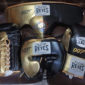 以下が含まれることがあります： Cleto Reyes 007ボクシングセット。黒と金のレースアップグローブ2つ、ヘッドギア、ファウルカップが含まれています。各アイテムにはCleto Reyesのロゴと金色の007エンブレムが施されています。ギアは黒と金のツートンカラーのデザインで構成されています。