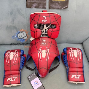 Op de afbeelding: Een set rode en blauwe boksuitrusting met een spinnenwebpatroon en witte spinnenemblemen. De set bevat een hoofdbeschermer en twee bokshandschoenen, allemaal voorzien van het woord FLY. Een smartphone toont het Punchwaveboxinggear-logo.