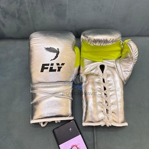 Puede incluir: Guantes de boxeo plateados con detalles amarillos y cordones blancos. Los guantes tienen la palabra "FLY" impresa en negro con un gr&aacute;fico de p&aacute;jaro. Un smartphone con un logotipo est&aacute; en primer plano.