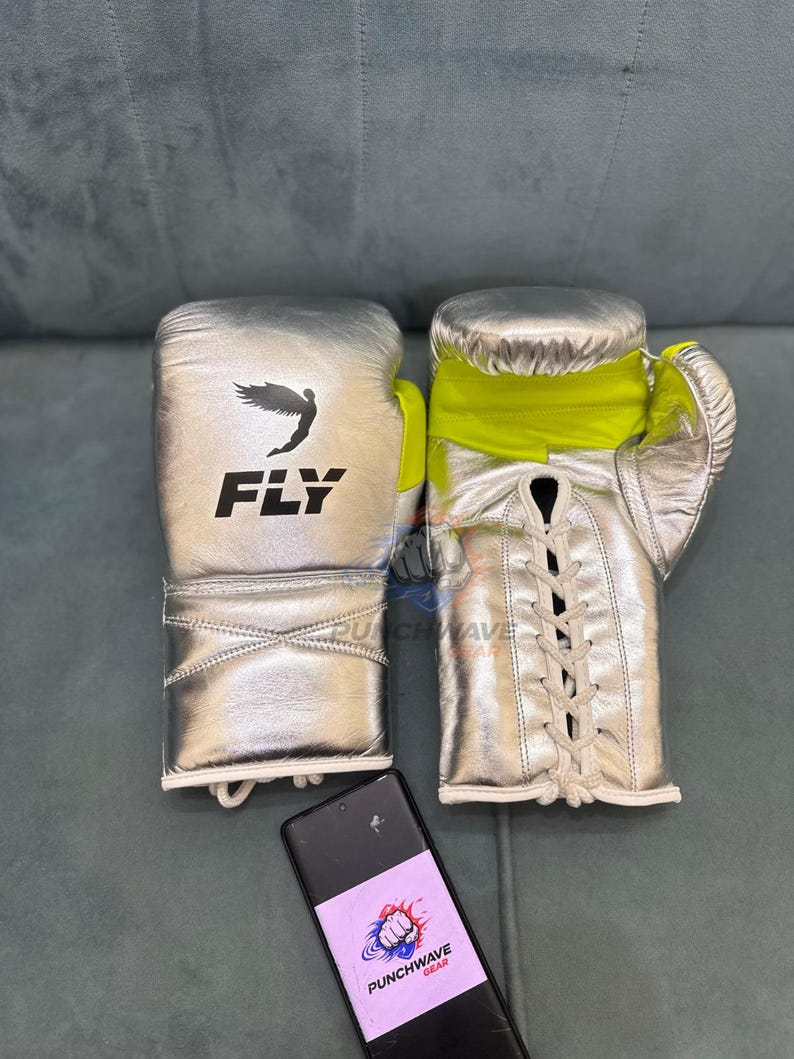 Puede incluir: Guantes de boxeo plateados con detalles amarillos y cordones blancos. Los guantes tienen la palabra "FLY" impresa en negro con un gr&aacute;fico de p&aacute;jaro. Un smartphone con un logotipo est&aacute; en primer plano.