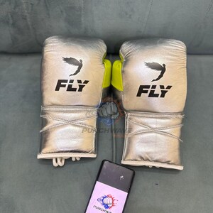 Puede incluir: Un par de guantes de boxeo plateados con la palabra "FLY" impresa en negro. Los guantes tienen un logotipo de p&aacute;jaro negro y detalles en amarillo ne&oacute;n. Un smartphone con el logotipo "PUNCHWAVE GEAR" est&aacute; en primer plano.