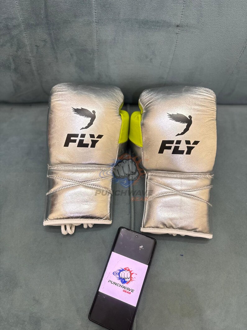 Puede incluir: Un par de guantes de boxeo plateados con la palabra "FLY" impresa en negro. Los guantes tienen un logotipo de p&aacute;jaro negro y detalles en amarillo ne&oacute;n. Un smartphone con el logotipo "PUNCHWAVE GEAR" est&aacute; en primer plano.