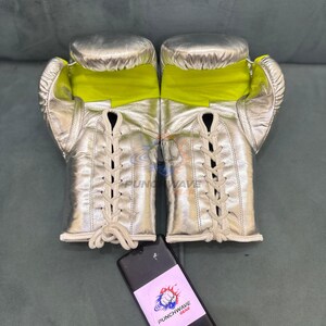 Puede incluir: Guantes de boxeo plateados con interiores amarillo ne&oacute;n y cordones color crema. Los guantes se exhiben sobre una superficie gris. Un tel&eacute;fono negro con una pegatina "PUNCHWAVE GEAR" est&aacute; en primer plano.