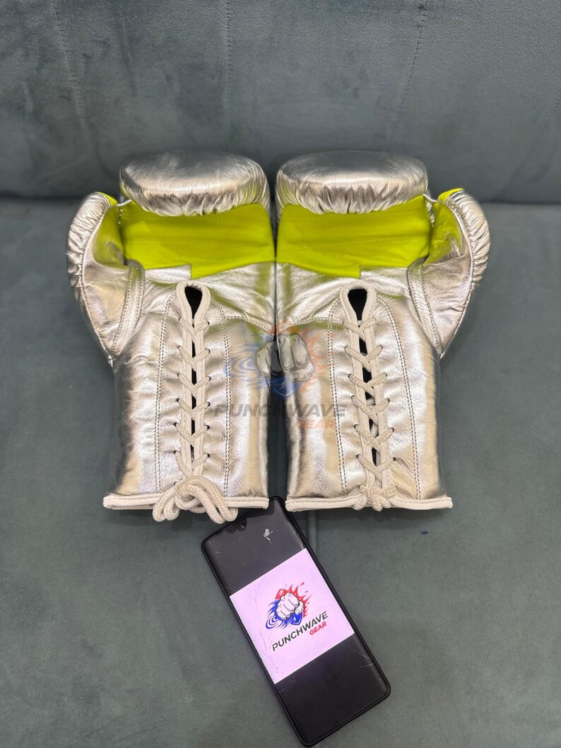 Puede incluir: Guantes de boxeo plateados con interiores amarillo ne&oacute;n y cordones color crema. Los guantes se exhiben sobre una superficie gris. Un tel&eacute;fono negro con una pegatina "PUNCHWAVE GEAR" est&aacute; en primer plano.
